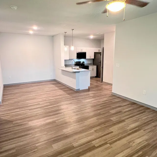 A spacious and modern apartment interior with light wooden flooring, a ceiling fan, and a kitchen area visible in the background.