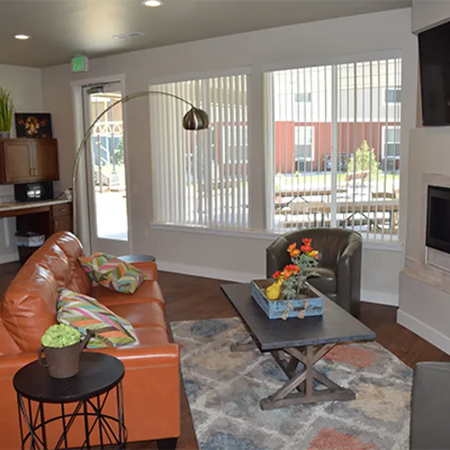 A modern living room featuring a brown leather couch with colorful pillows, a round table, green armchairs, a coffee table, and a wall-mounted television. There are large windows with blinds allowing natural light, and a decorative centerpiece on the coffee table.