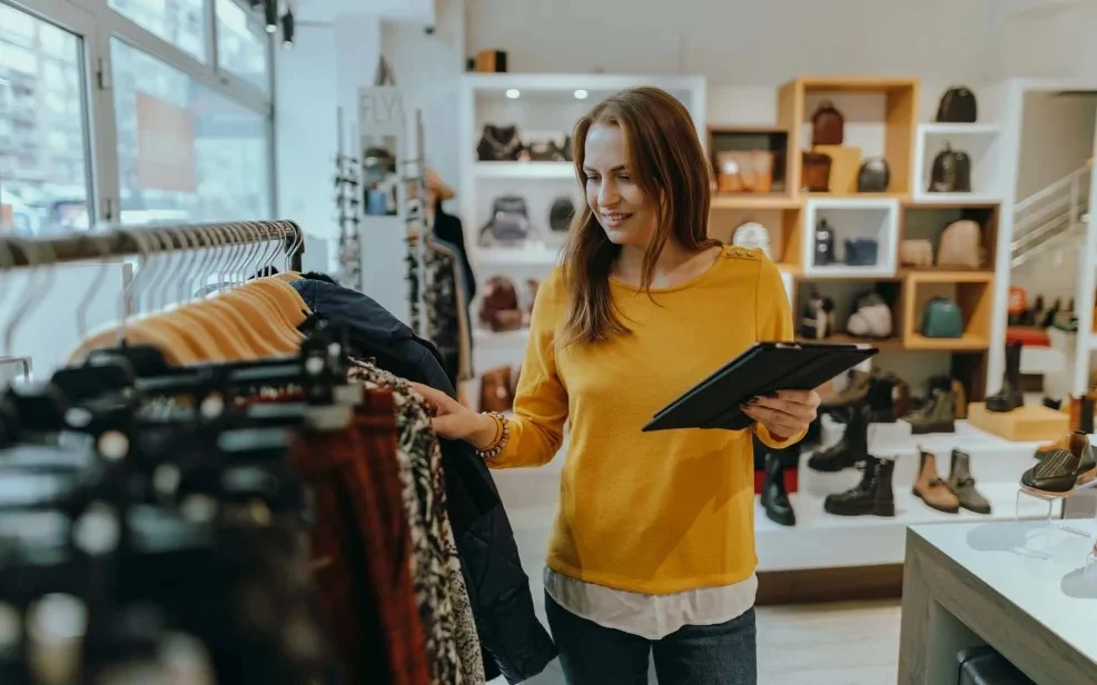 A woman browsing clothing racks in a boutique, smiling while holding a tablet. The store is brightly lit with shelves displaying various bags and shoes in the background.