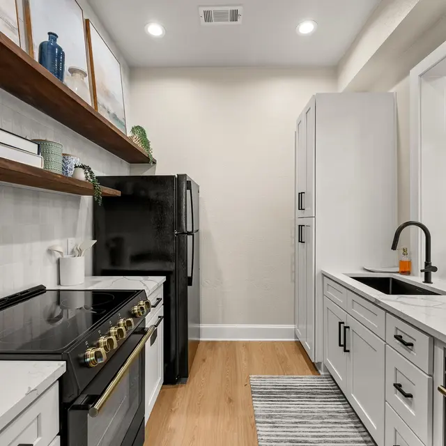 A modern kitchen featuring a black stove, white cabinetry, and open wooden shelves holding decorative items. Light-colored walls and a hardwood floor enhance the spacious feel of the kitchen.