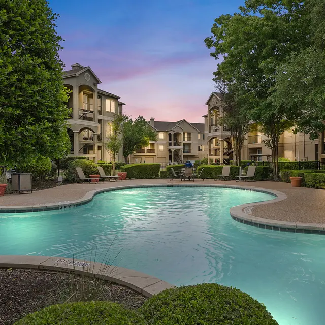 A beautiful swimming pool surrounded by lush greenery and apartment buildings at dusk, with a pastel sky in the background.