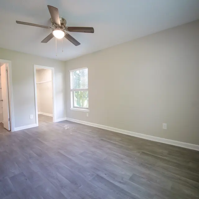 A spacious empty room with gray laminate flooring, a ceiling fan, and a window letting in natural light. A doorway leads to a closet or bathroom area.