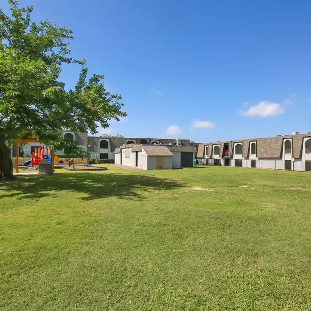 A spacious grassy area featuring a playground and surrounding apartment buildings under a clear blue sky.