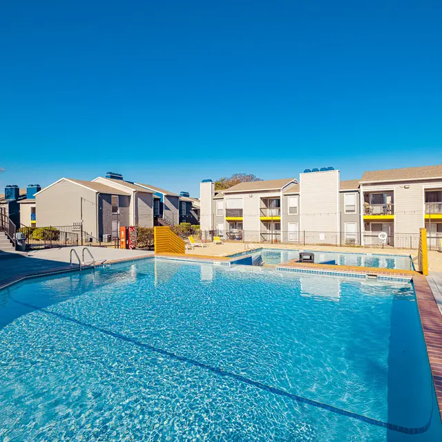 A clear blue swimming pool surrounded by a sandy area and apartment buildings in the background.