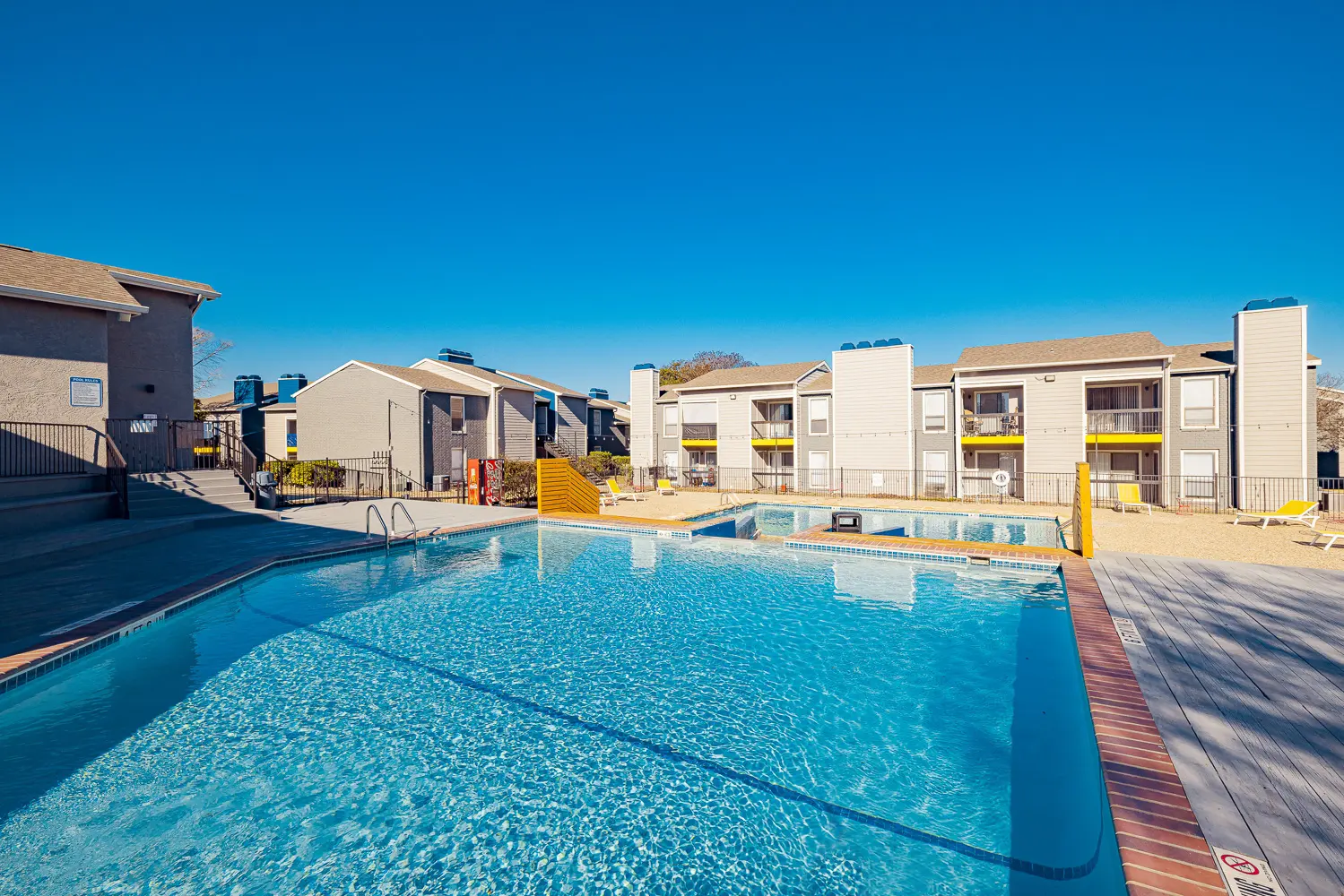 A clear blue swimming pool surrounded by a sandy area and apartment buildings in the background.