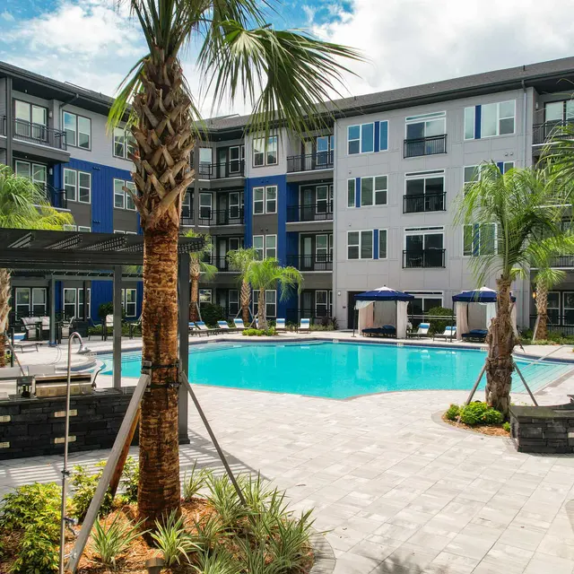 A modern apartment complex featuring a swimming pool surrounded by palm trees and lounge chairs.