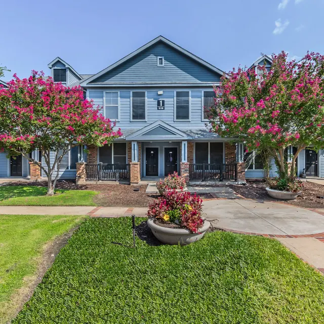 A residential apartment complex with two stories, featuring blue siding and landscaped front yard. Decorative pink flowering trees are on both sides of the entrance, complementing neatly trimmed green grass and flowerbeds.