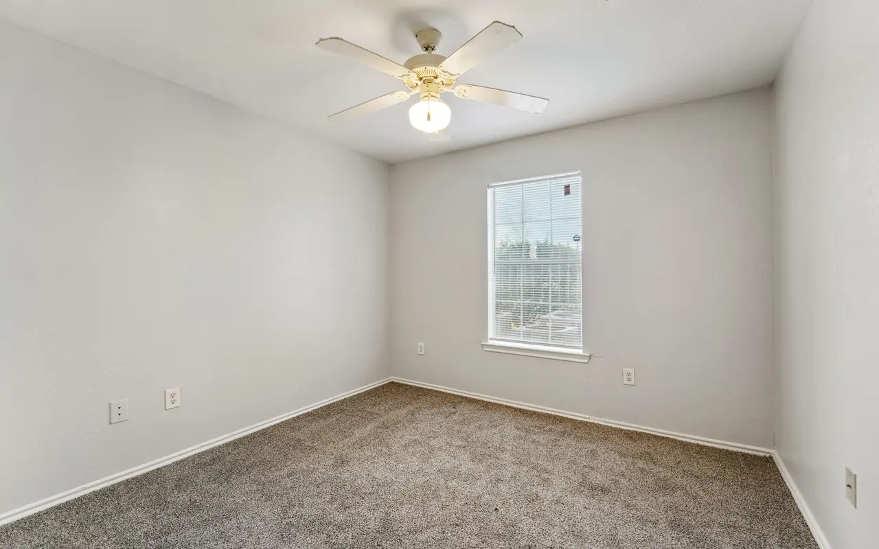 A spacious, empty bedroom featuring light gray walls and a light brown carpet. A ceiling fan hangs from the ceiling, and there is a single window allowing natural light to enter the room.