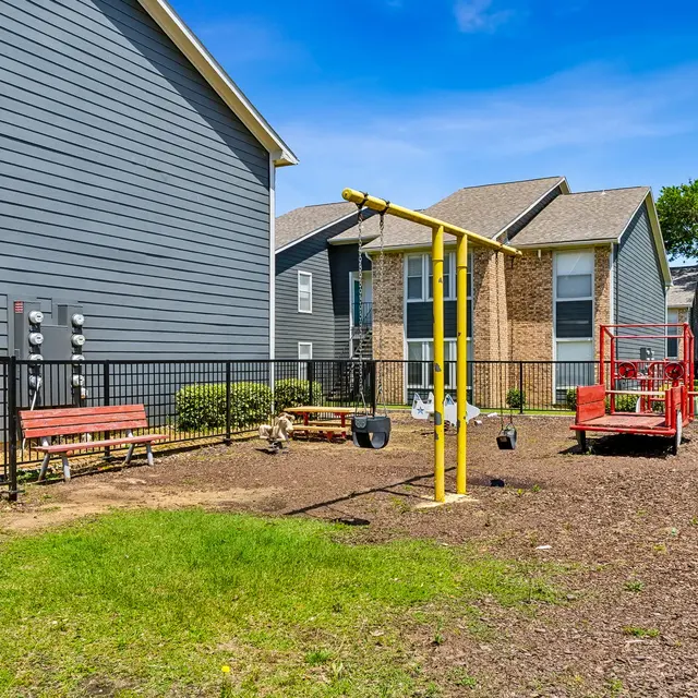 A fenced playground area with swings and a red play structure, surrounded by grassy patches and benches. The setting features a mix of gray and tan buildings in the background under a clear blue sky.