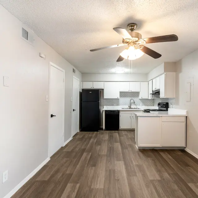 A modern kitchen with a ceiling fan, laminate flooring, and white cabinetry. There is a black refrigerator and stainless steel sink visible, with a window allowing natural light in.