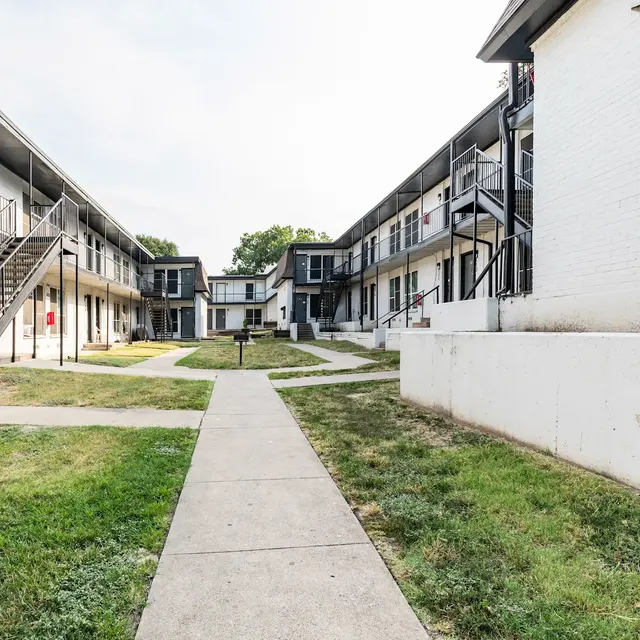 A view of a courtyard in an apartment complex, featuring a pathway surrounded by two-story buildings with balconies. The area is green with grass on either side of the walkway.