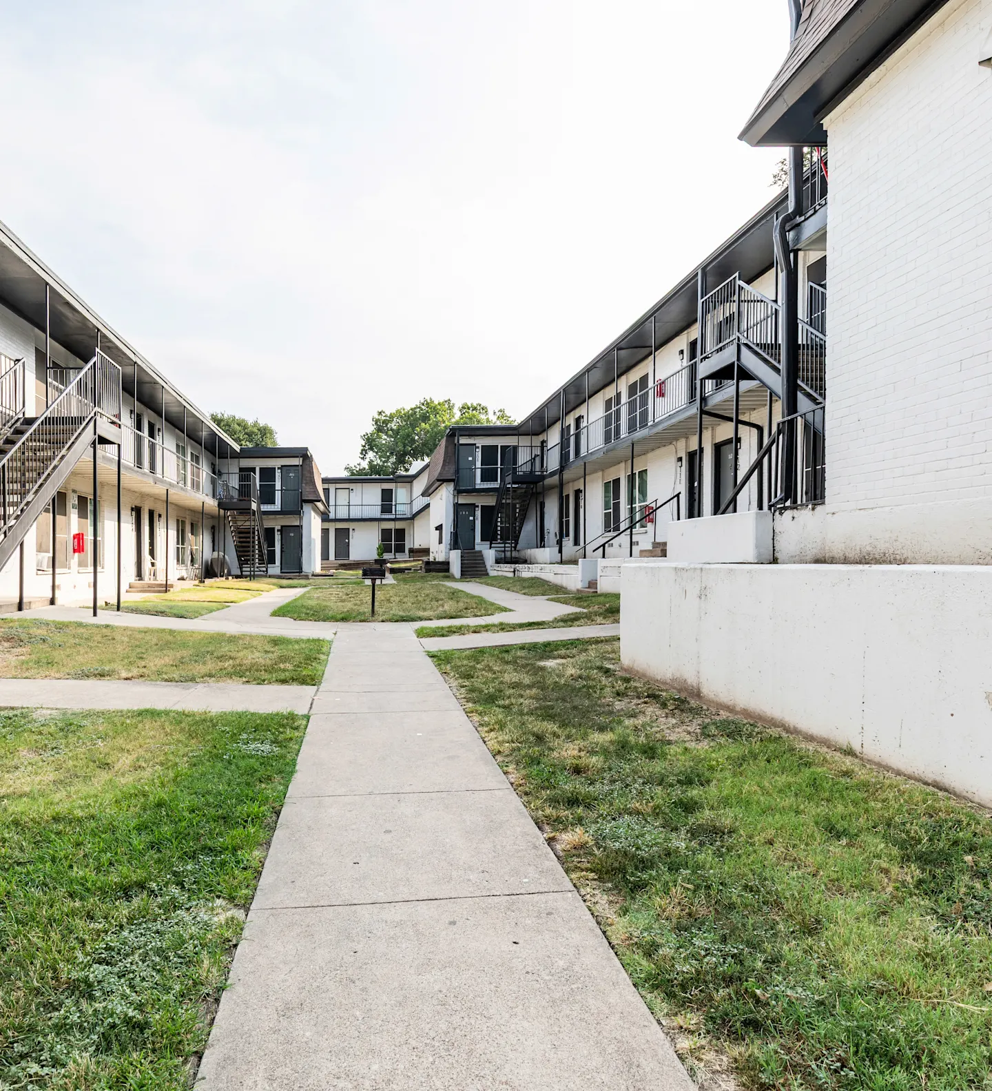 Apartment Complex Courtyard A view of a courtyard in an apartment complex, featuring a pathway surrounded by two-story buildings with balconies. The area is green with grass on either side of the walkway.