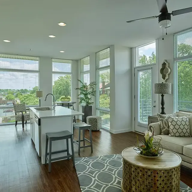 A bright, modern open concept kitchen and living area with large windows and views of greenery outside. The kitchen features a white island, stainless steel appliances, and a cozy living area with a couch and decorative pillows.