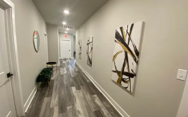 A modern hallway featuring light gray walls, wooden flooring, and contemporary abstract art pieces. A small green bench is visible along the side.