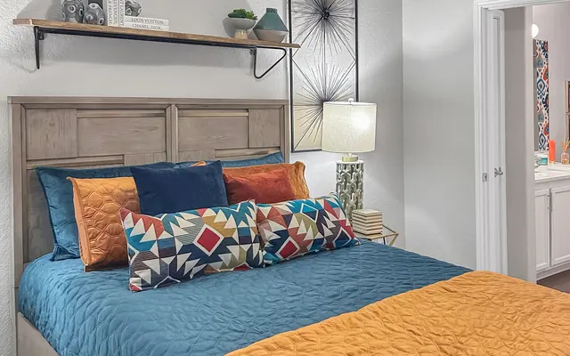 A stylish bedroom featuring a bed with teal and orange bedding, accent pillows in geometric patterns, a bedside lamp, and a shelf above the headboard with decorative items.
