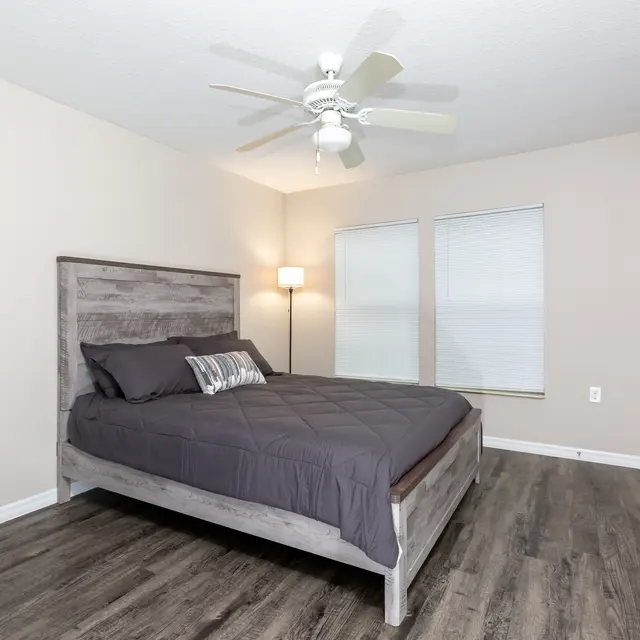 A modern bedroom featuring a gray platform bed with a quilted dark bedding, a bedside lamp, and two windows.