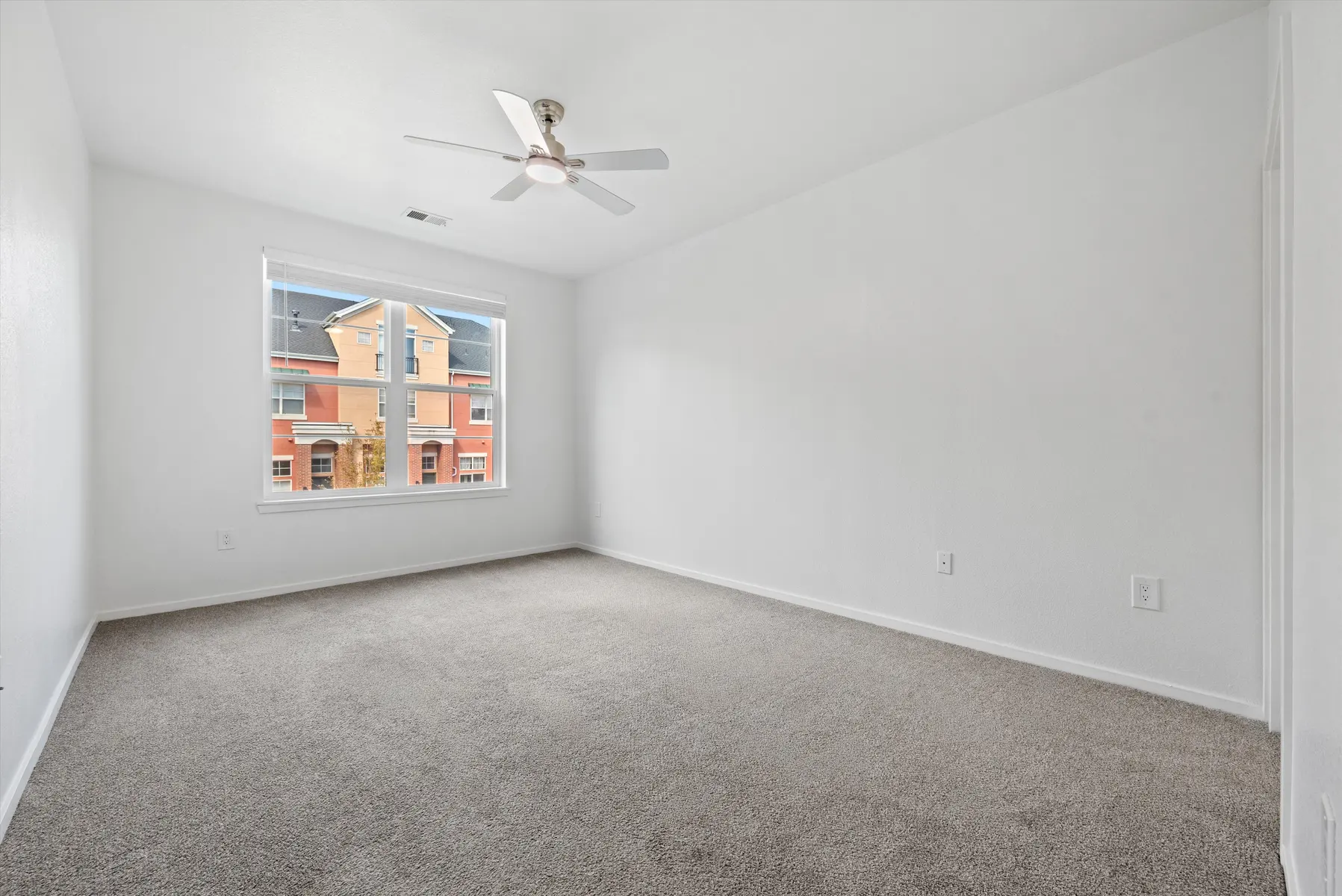 Empty bedroom with gray carpet and white walls, featuring a ceiling fan and a window showing a colorful exterior view.
