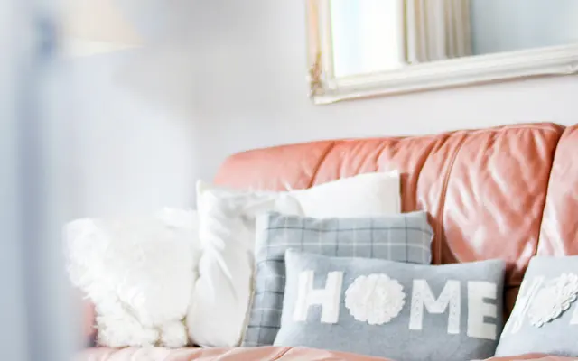 A cozy living room with a salmon-colored couch adorned with decorative pillows, including one that says 'HOME'. A light blue ottoman sits in front of the couch, and there's a mirror on the wall reflecting natural light.