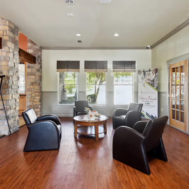Cozy Lobby Interior A cozy lobby area featuring a round table with a flower arrangement and four black chairs arranged around it. Natural light filters in through several windows, and there is a stone accent wall on one side. A banner and framed photo are displayed nearby.