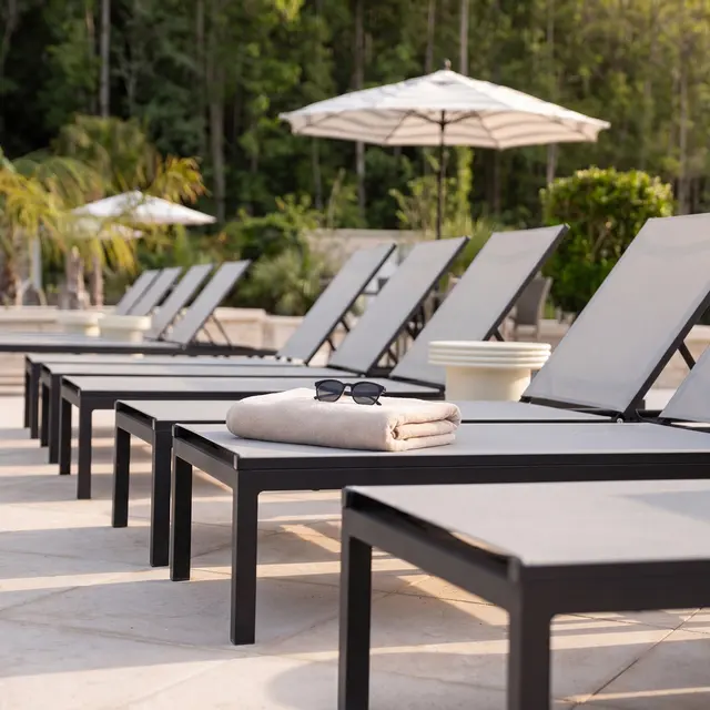 Relaxing Poolside Loungers A row of modern poolside loungers featuring gray cushions, with a towel and sunglasses placed on one of them, under a bright sunny sky and surrounded by greenery.