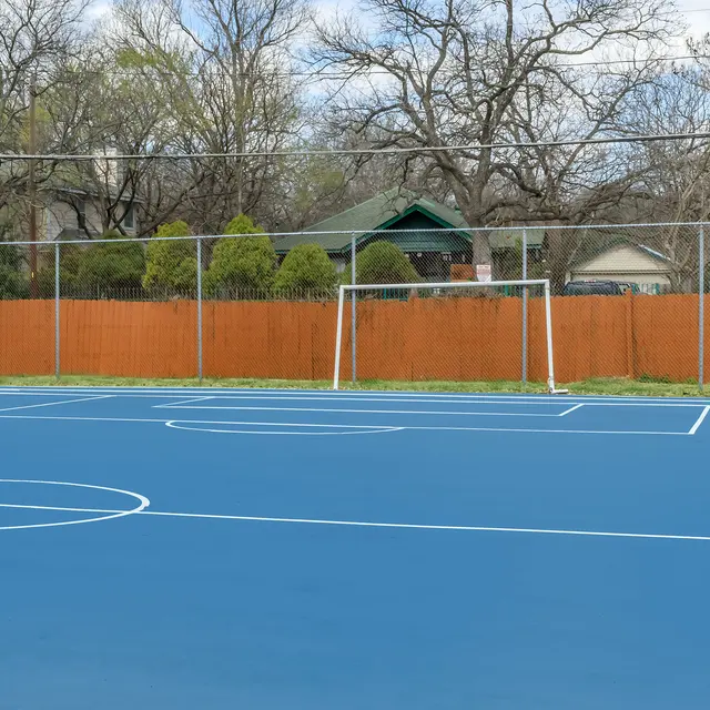 A newly painted blue soccer field with white markings, featuring a goal at one end and a fence in the background. Trees and a green house are visible behind the fence.