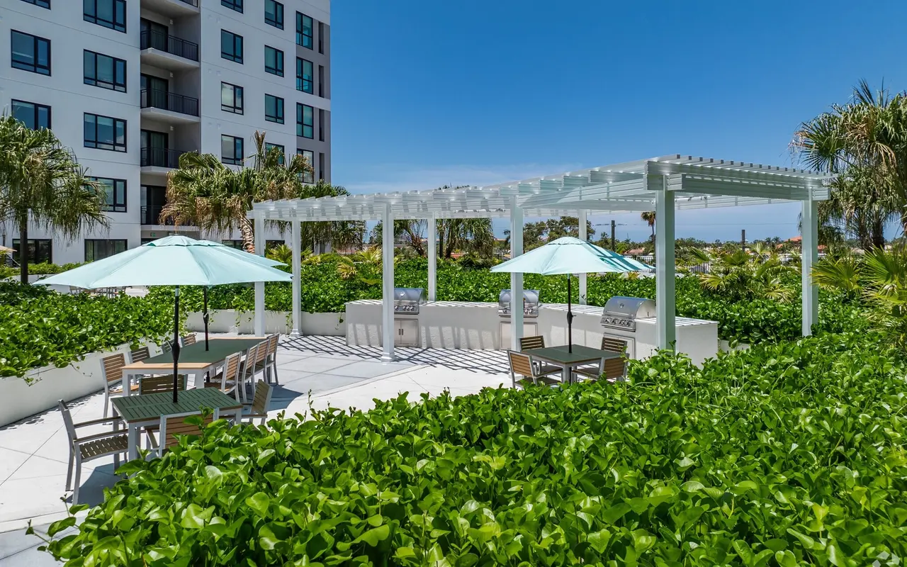 A patio area surrounded by lush greenery with white pergolas and umbrellas. The background features a modern building and blue sky.