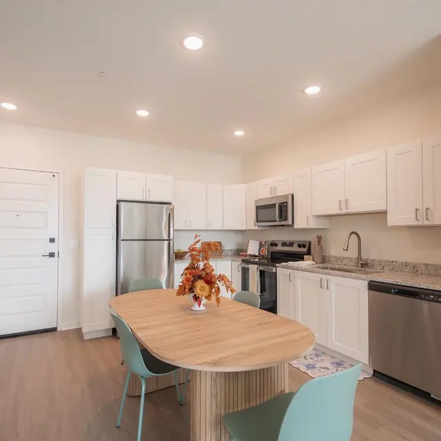 A modern kitchen and dining area featuring a wooden table with a bouquet of flowers, light-colored cabinetry, stainless steel appliances, and a neutral color palette.