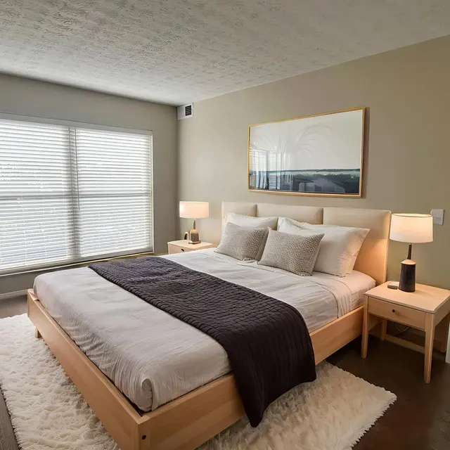 A modern bedroom featuring a large bed with light-colored linens and a dark throw blanket, two bedside lamps, a white dresser, and a plant nearby. Large windows allow natural light to brighten the space.