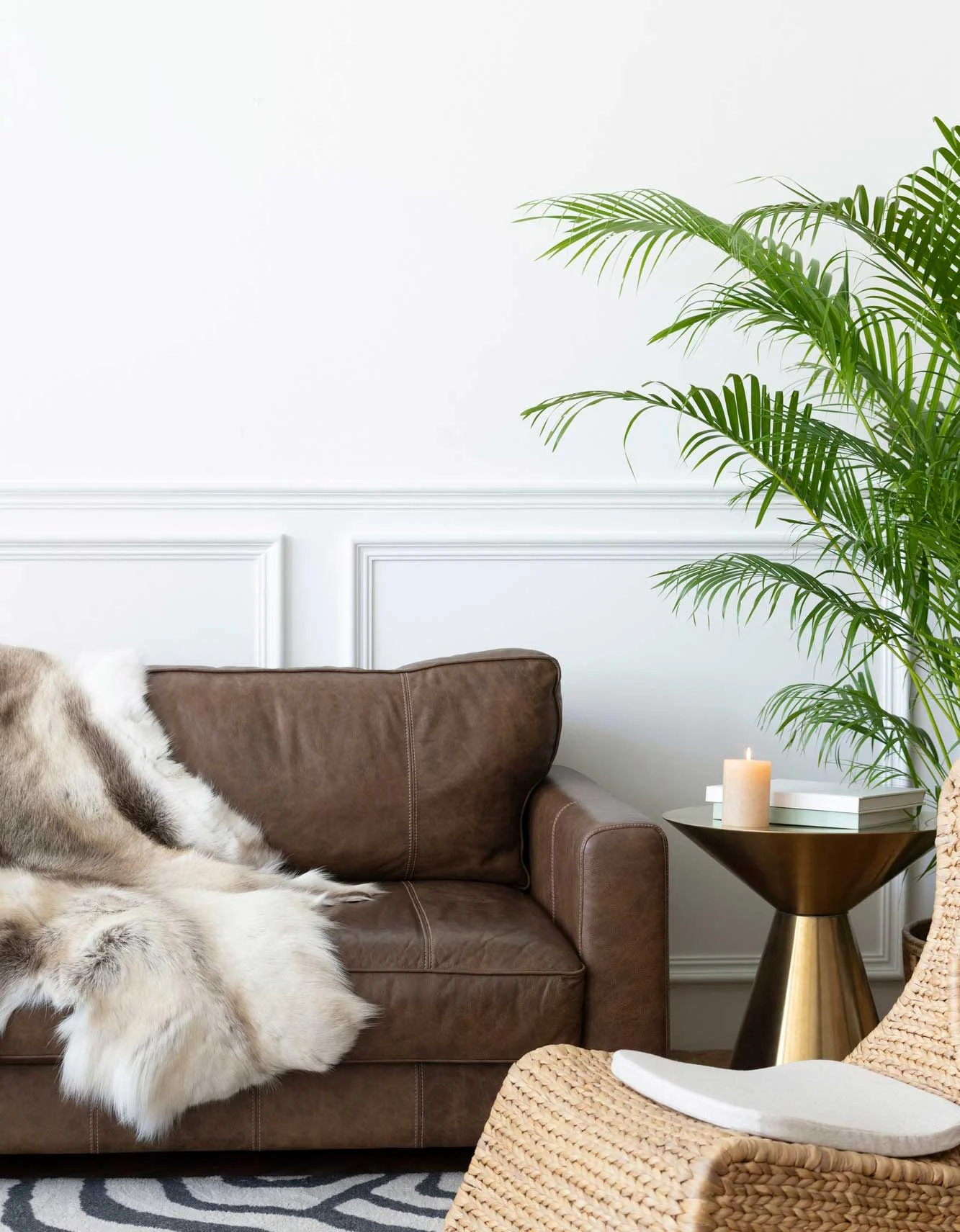 A cozy living room featuring a brown leather sofa with a fur throw, a small round table with a candle, decorative plant, and a woven chair on a patterned rug.