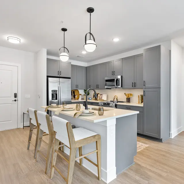 A modern kitchen featuring gray cabinetry, stainless steel appliances, and a white island with seating. The floor is light wood, and there are pendant lights hanging above the island. A doorway leads to a bathroom area, indicating a spacious layout.