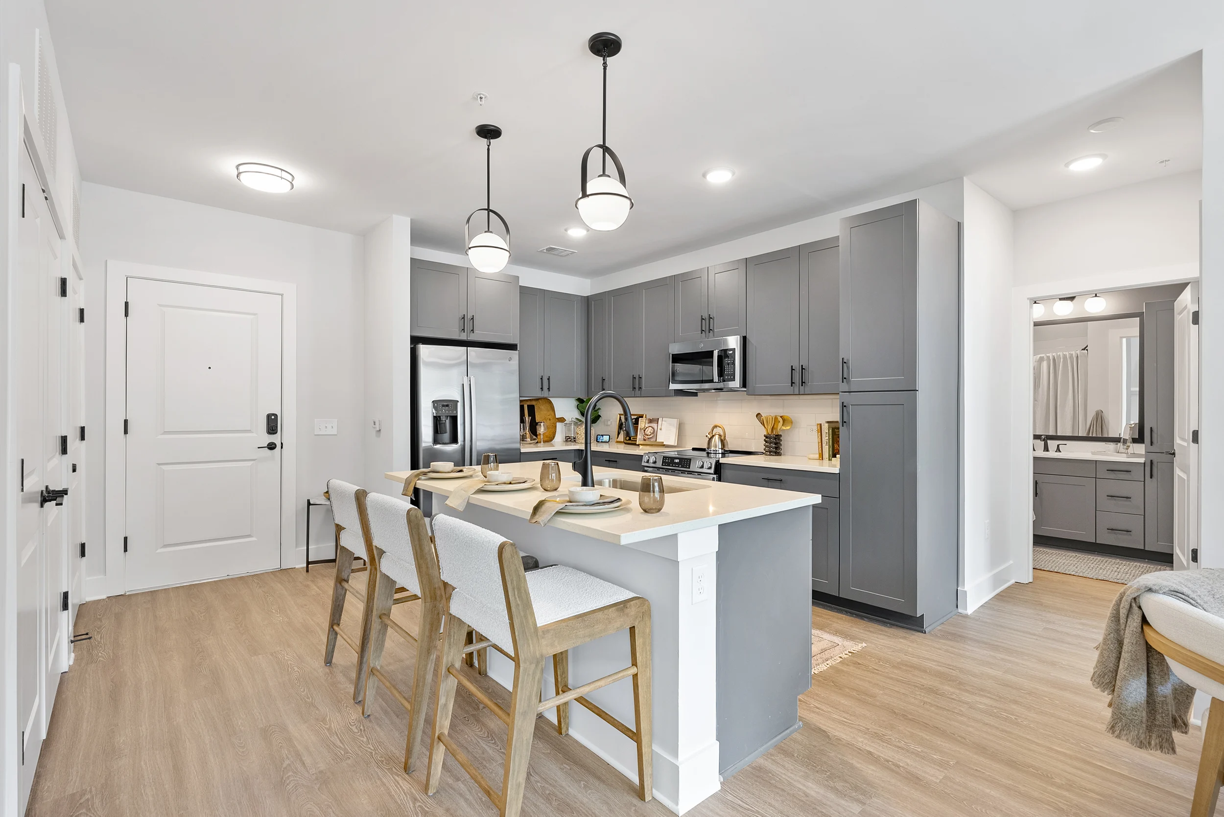 Stylish Modern Kitchen Interior A modern kitchen featuring gray cabinetry, stainless steel appliances, and a white island with seating. The floor is light wood, and there are pendant lights hanging above the island. A doorway leads to a bathroom area, indicating a spacious layout.
