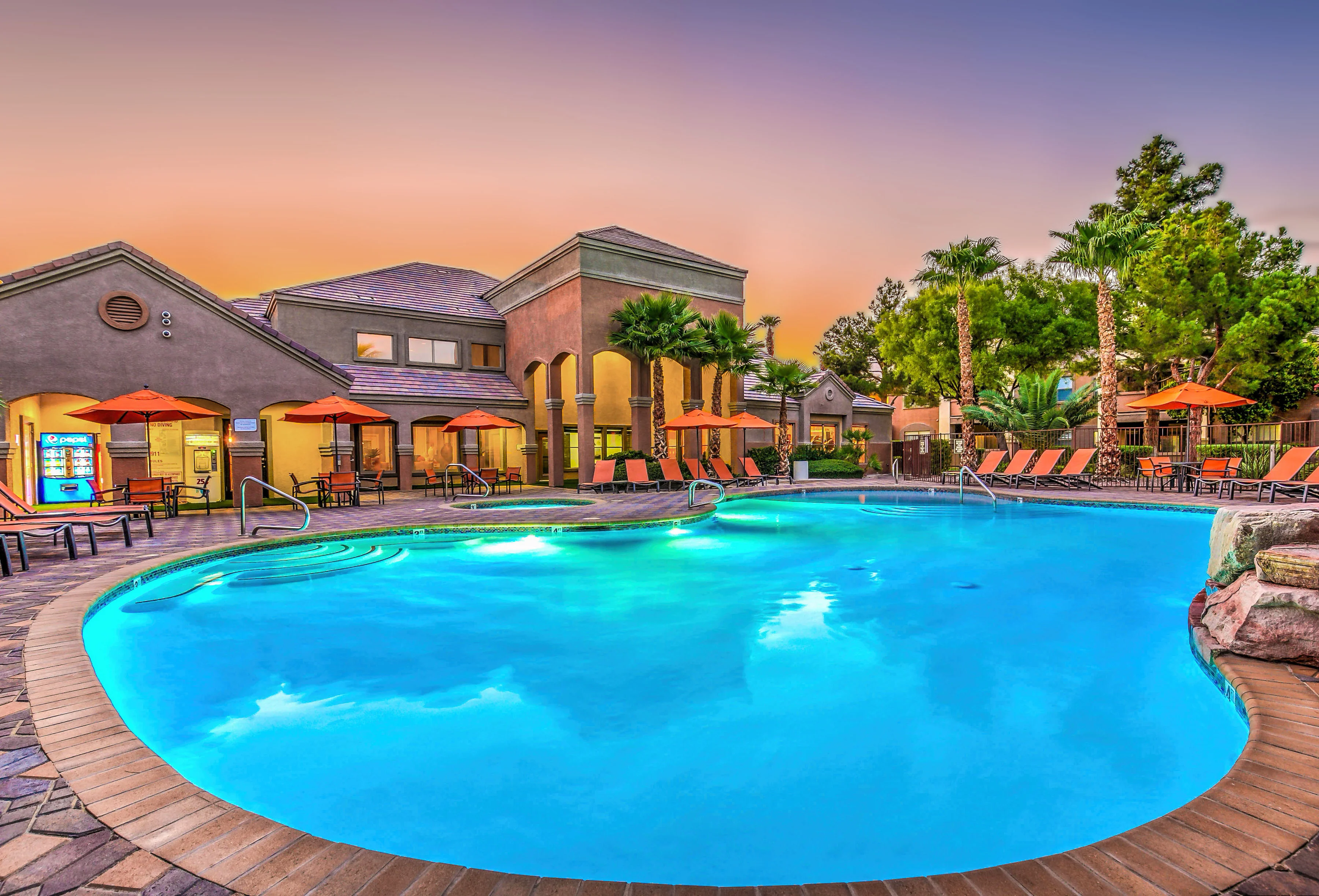An inviting outdoor pool area with lounge chairs and umbrellas, surrounded by palm trees and a residential building, under a colorful sunset sky.