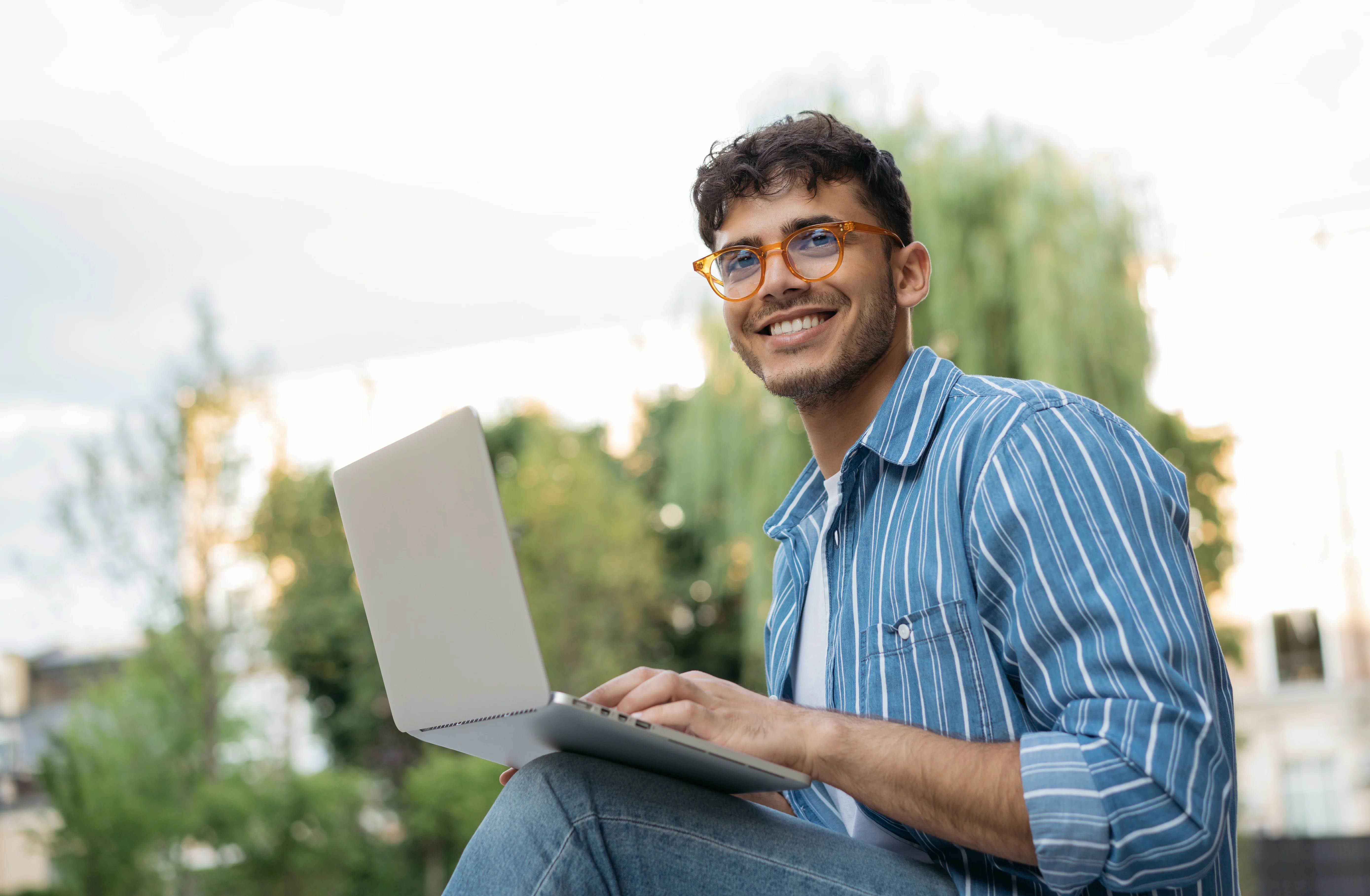 A young man sitting outdoors, smiling while using a laptop. He is wearing glasses and a blue striped shirt, with greenery in the background.