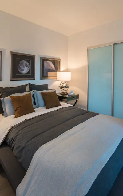A modern bedroom featuring a queen-sized bed with gray and white bedding, illuminated by two stylish bedside lamps. The walls have framed celestial images, and a sliding glass door is visible in the corner. There is a small decorative plant and a side table with an intricate design.