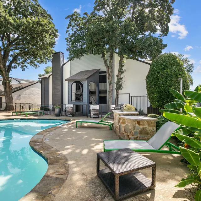 A swimming pool area with lounge chairs and surrounding green foliage. The pool is clear and reflects the blue sky, with a building visible in the background.