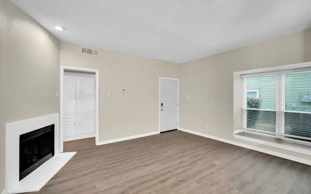 A spacious living room with light-colored walls and a wood floor. There is a fireplace on the left, a door leading outside in the center, and a large window on the right with a window seat underneath.