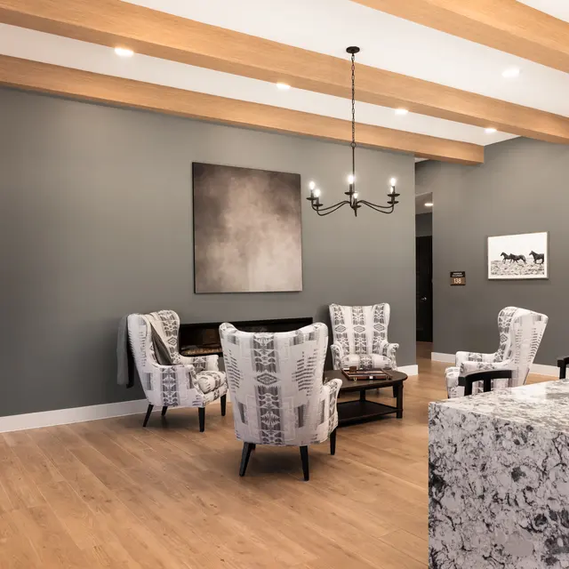 A modern living room featuring two patterned armchairs, a wooden table, and a gray accent wall with a piece of art. There are wooden beams on the ceiling and a textured wall made of stacked wood in the background.