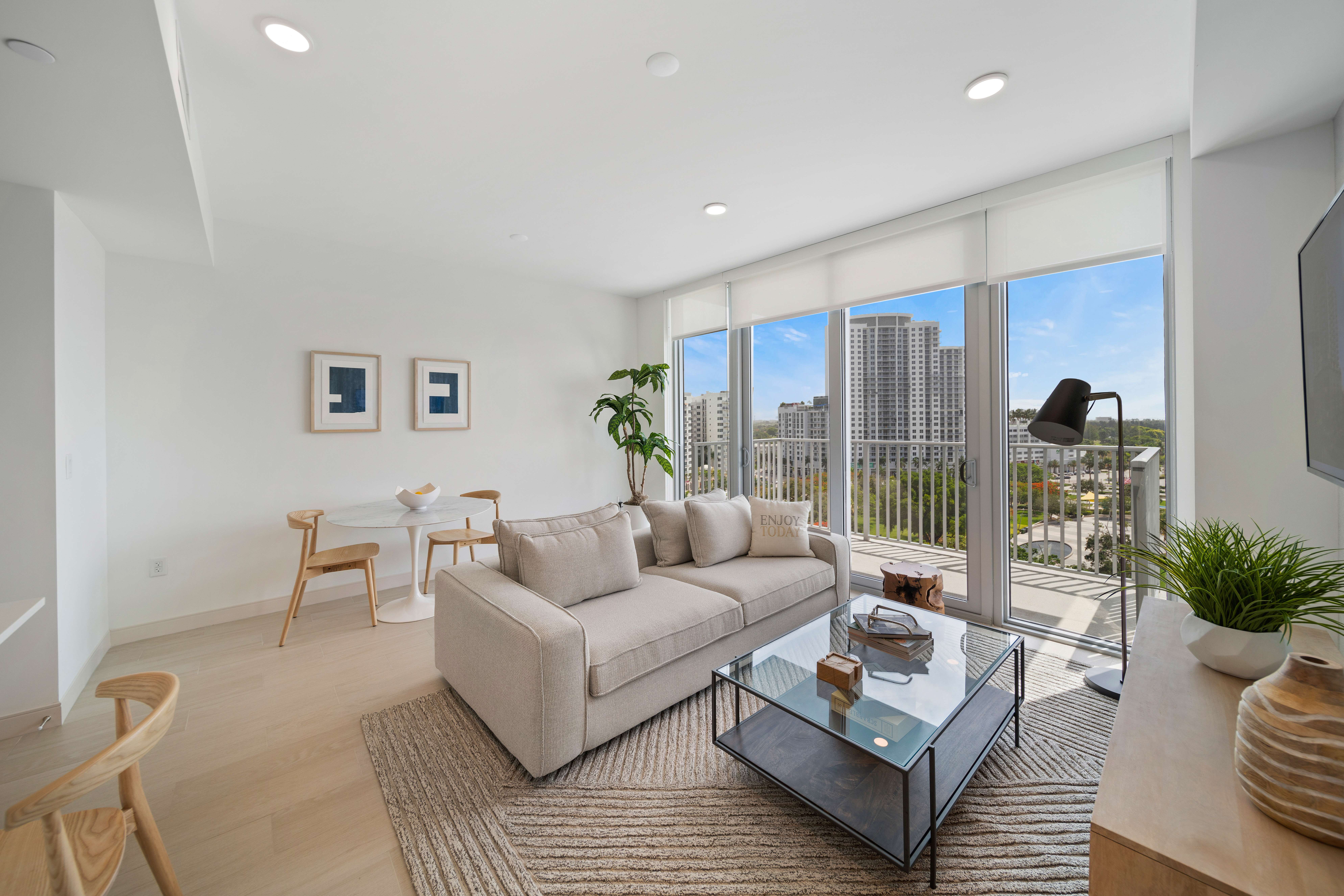 A modern living room featuring a gray sofa, glass coffee table, and large windows with a city view. The room has light-colored walls and wooden flooring.