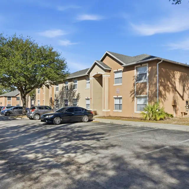 A beige apartment complex with parked cars in front, surrounded by trees and blue sky.