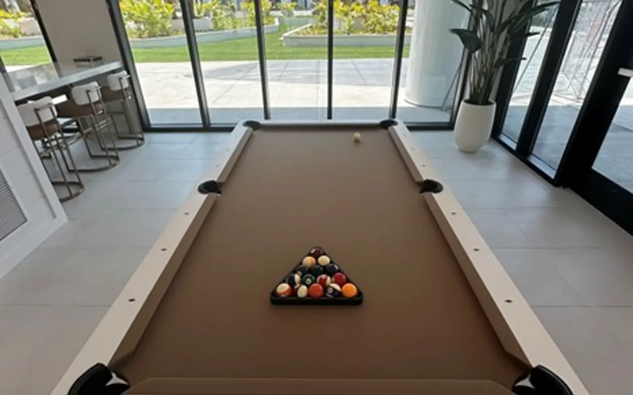 A modern pool table with a triangular rack of colorful balls set up for a game. The space is well-lit with large windows providing views of a green outdoor area. Minimalistic furniture and a pendant light are visible in the background.