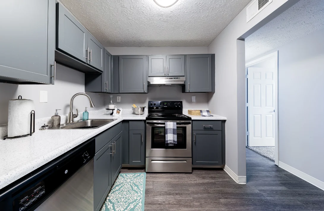 A modern kitchen featuring gray cabinets, stainless steel appliances, and a white countertop. A teal mat is on the floor, and a doorway leads to another space.