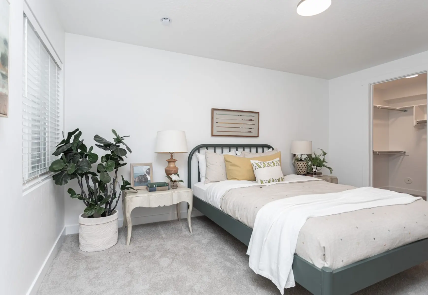 Modern Bedroom Interior A modern bedroom featuring a bed with gray and white bedding, two bedside lamps, and a potted plant. The room has a light-colored carpet and white walls, with a window partially covered by blinds.