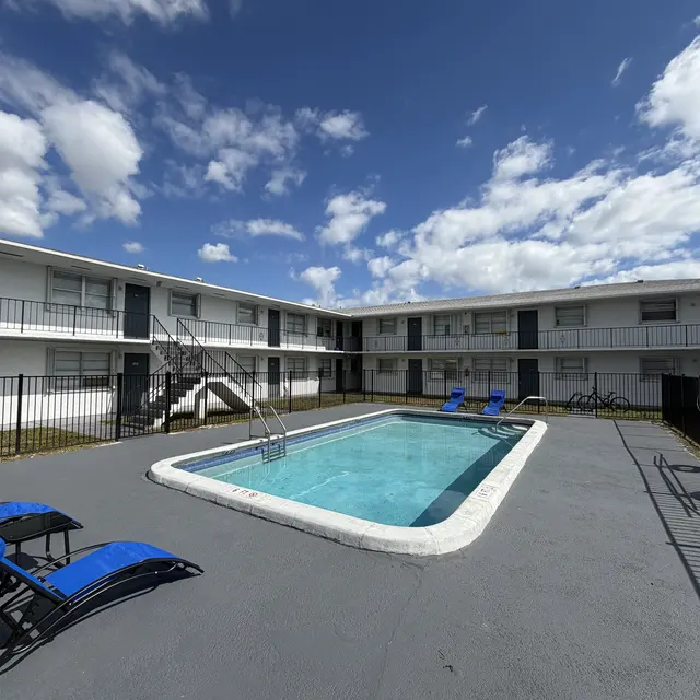 A view of a pool area surrounded by a low-rise apartment building. There are blue lounge chairs by the pool, and the sky is partly cloudy with a few visible clouds.