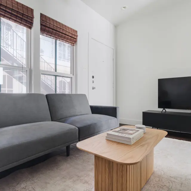 A modern living room featuring a gray sofa, a wooden coffee table, and a television on a low cabinet. Bright natural light enters through wide windows with wooden blinds.
