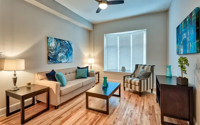 Modern Living Room Interior Design A cozy living room featuring a beige sofa with blue cushions, a striped armchair, and a wooden coffee table. The room is well-lit with a lamp, has hardwood flooring, and decorative wall art in blue tones.