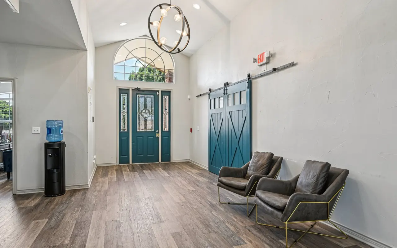 A modern entryway featuring two dark leather chairs, a decorative chandelier, and a large window with an arch above the double doors. There is also a water dispenser in the corner.
