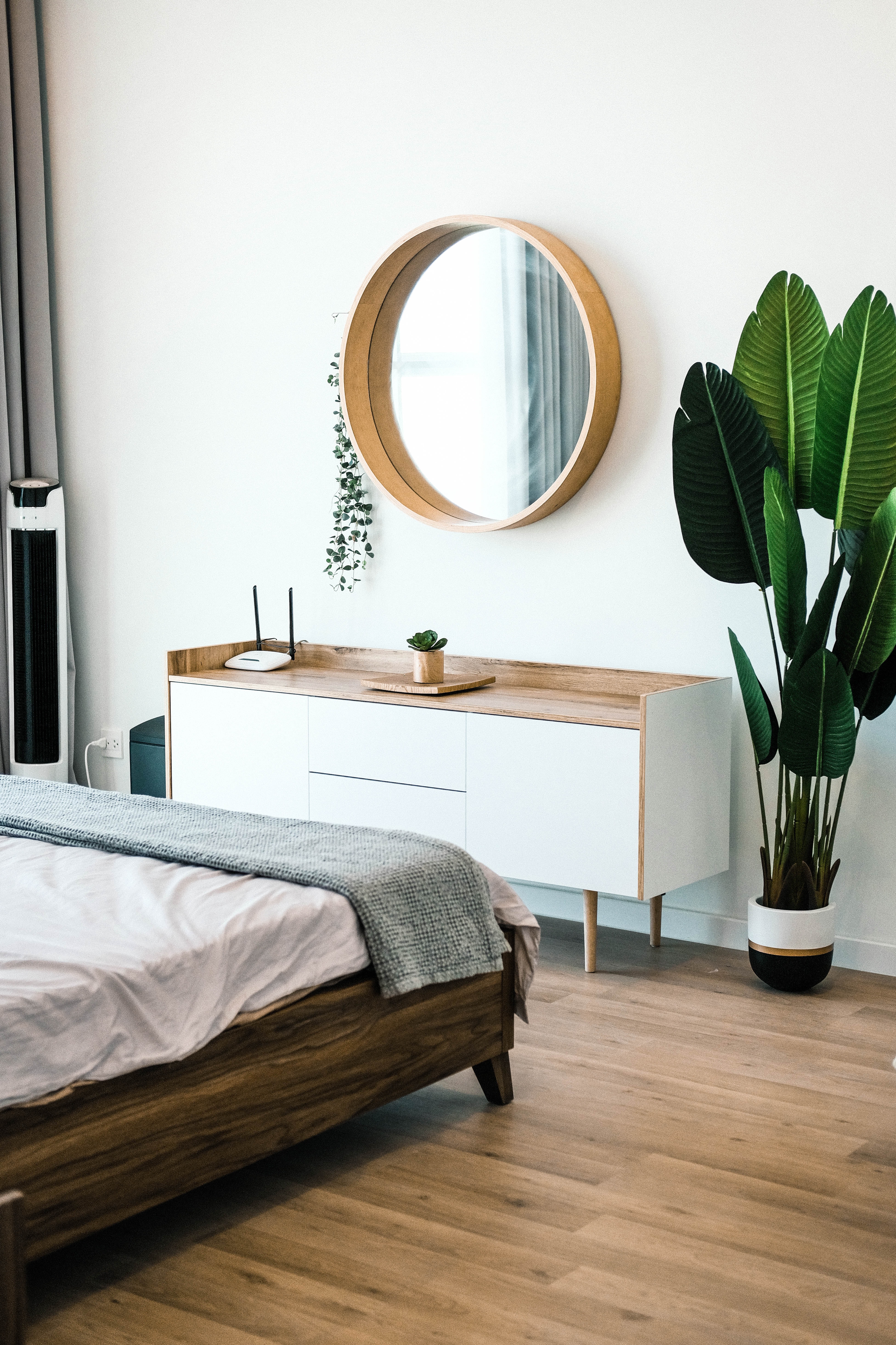 A modern bedroom featuring a wooden bed, a white cabinet with a circular mirror above it, and a large potted plant.