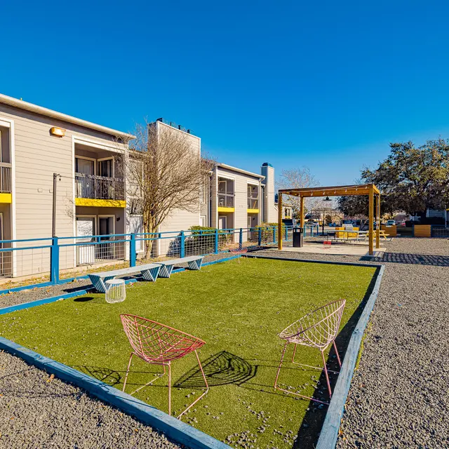 A playground area surrounded by apartments, featuring green grass, gravel pathways, and colorful chairs.