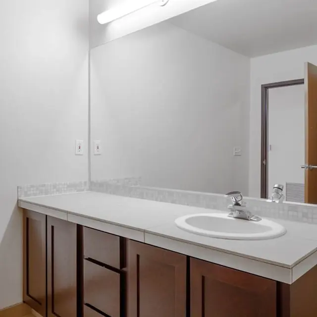 A clean and modern bathroom featuring a simple countertop with a sink, brown cabinetry below, and a large mirror. The walls are painted white, and there is a door leading to another room in the background.