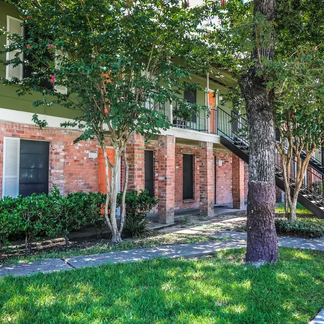 Exterior view of an apartment complex featuring brick and green siding, trees, and grassy areas.